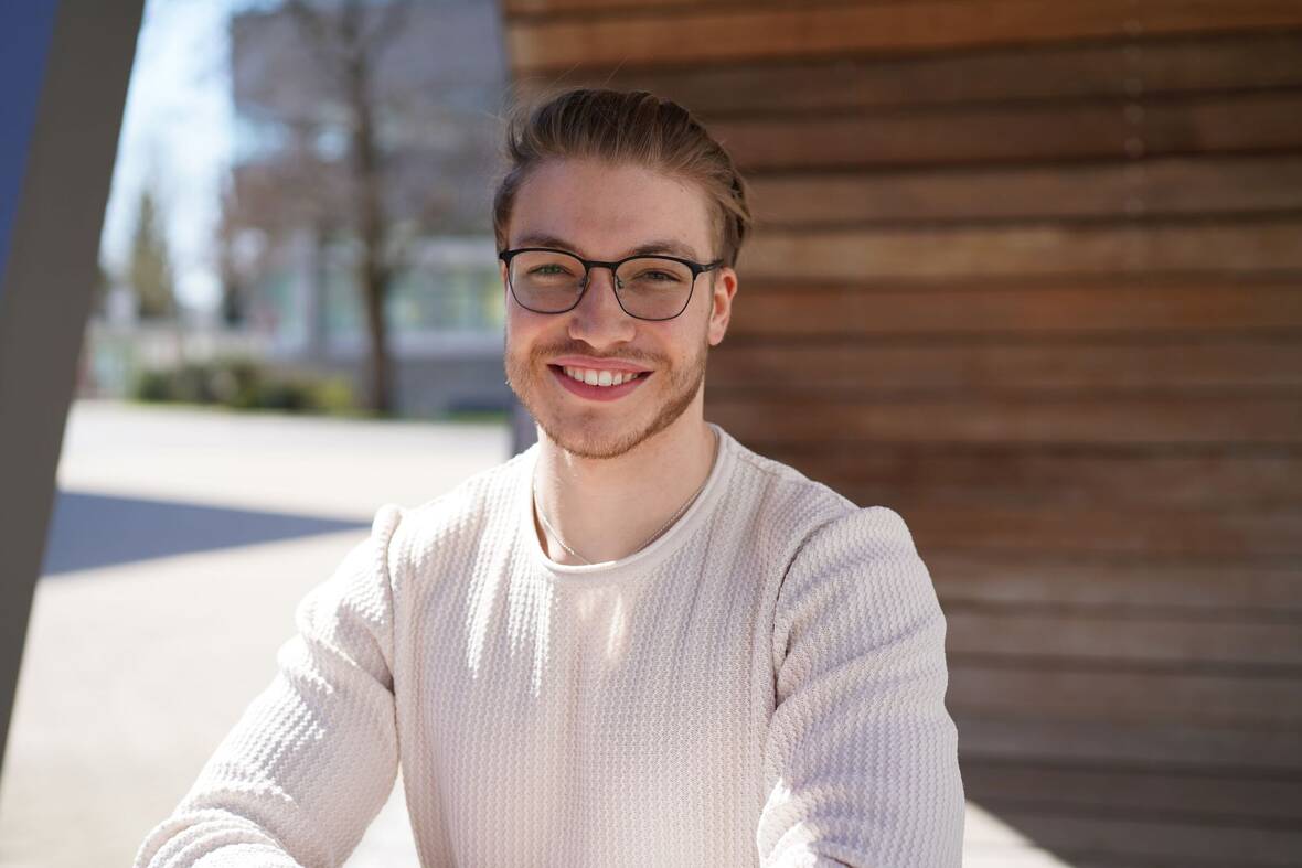 Ein junger Mann sitzt auf dem OTTO Campus. Hinter ihm sind Holzpaneele zu sehen. Er trägt eine Brille, hat kurze Haare und guckt freundlich in die Kamera