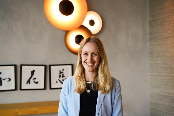 Lulu Eine Frau mit langen Haaren lächelt direkt zur Kamera. An der Wand im Hintergrund hängen moderne, kreisförmige Lampen und drei Bilder mit kalligraphierten japanischen Schriftzeichen.