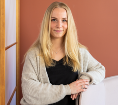 Eine junge Frau mit langen Haaren lehnt mit den Armen auf einen Sessel, während sie freundlich zur Kamera guckt