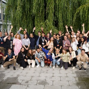 Auf einem gepflasterten Platz stehen die fast 50 neuen Azubis und dualen Student:innen. Sie sind in Reihen aufgestellt und und heben die Arme für das Foto. Hinter ihnen steht eine Weide und an der Seite ist ein Gebäude vom OTTO Campus zu erkennen.