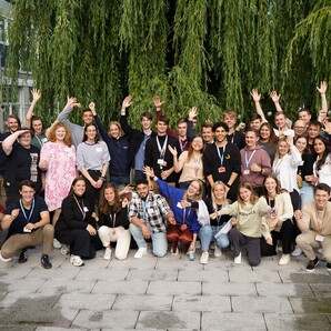 Auf einem gepflasterten Platz stehen die fast 50 neuen Azubis und dualen Student:innen. Sie sind in Reihen aufgestellt und und heben die Arme für das Foto. Hinter ihnen steht eine Weide und an der Seite ist ein Gebäude vom OTTO Campus zu erkennen.