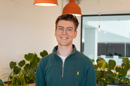 Autorenbild des jungen Techblog-Autoren René in einem dunkelgrünen Pullover mit Brille. Der Autor lächelt freundlich und befindet sich in einem Raum mit Pflanzen und orangen Hängelampen. Der Autor befindet sich im OTTO-Headquater.