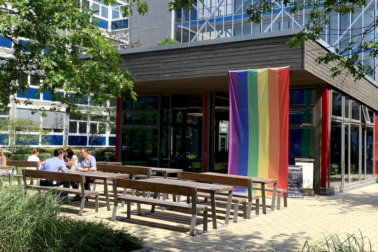 Auf dem Boulevard sind mehrere Bänke mit Tischen zu sehen, wo Mitarbeitende von OTTO sitzen. Im Hintergrund ist das kleine Gebäude des Bistros zu sehen, an dem die bunte LGTBQ-Flagge hängt. Drum herum sind mehrere Pflanzen.