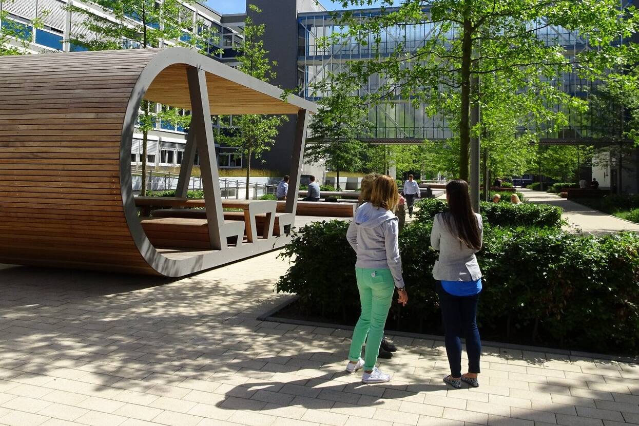 Neben den gepflasterten Wegen des Boulevards gibt es Grünflächen, auf denen Bäume und Büsche wachsen. Auf dem Boulevard steht auch eine moderne Konstruktion aus Holzbrettern und Stahl, die als überdachte Sitzgelegenheit dient.
