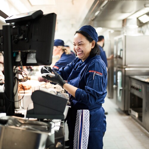 Unsere Köchin lacht begeistert auf ihren Bildschirm während sie in der Küche steht und die anderen Kollegen und Kolleginnen zu bedienen in der Kantine.
