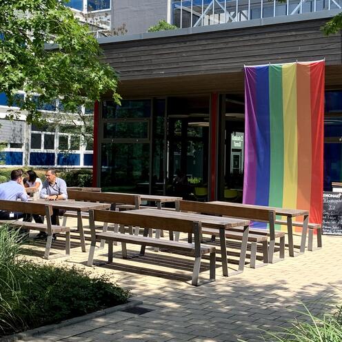 Auf dem Boulevard sind mehrere Bänke mit Tischen zu sehen, wo Mitarbeitende von OTTO sitzen. Im Hintergrund ist das kleine Gebäude des Bistros zu sehen, an dem die bunte LGTBQ-Flagge hängt. Drum herum sind mehrere Pflanzen.