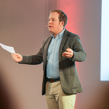 Ein Kollege steht vor einem Podium, redet in ein Mikrofon und hält in der anderen Hand einen Zettel.