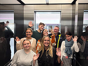 Leute stehen im Fahrstuhl und winken in die Kamera