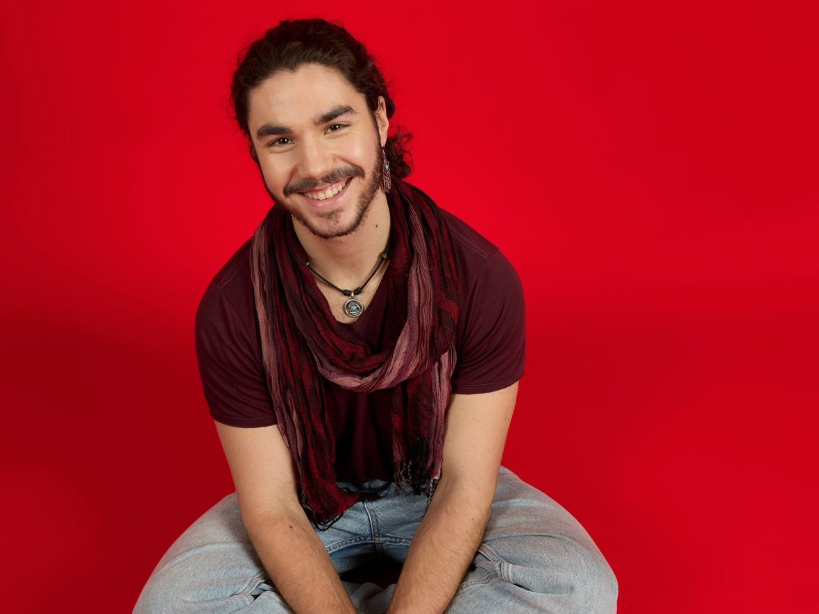 Unser Azubi sitzt im Schneidersitz auf dem Boden vor einem roten Hintergrund und lächelt in die Kamera. 