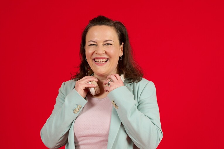 Unsere Technical Writerin lächelt in die Kamera Unsere Technical Writerin steht vor einem roten Hintergrund. Sie hat Kopfhörer um den Hals und hält diese mit den Händen fest. Unsere Kollegin lächelt freundlich in die Kamera.