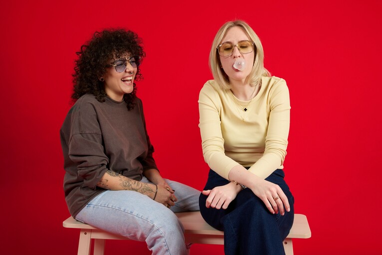 Unsere Kolleginnen sitzen aufm Stuhl Unsere Kolleginnen sitzen vor einem roten Hintergrund auf einer Bank. Die eine lacht die andere an, da sie eine Kaugummi Blase macht. Beide tragen Sonnenbrillen.
