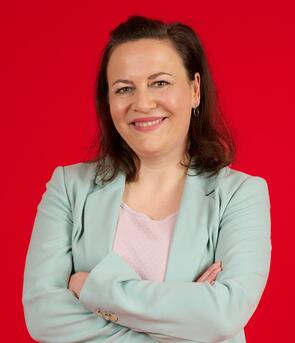 Unsere Technical Writerin steht vor einem roten Hintergrund. Sie verschränkt ihre Arme und lächelt freundlich in die Kamera. 