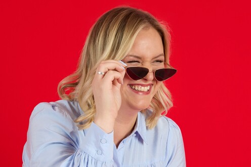 HR Managerin aus Digital Learning trägt eine Sonnenbrille und steht vor rotem Hintergrund. Sie greift mit ihrer linken Hand nach der Sonnenbrille und lacht nach rechts aus dem Bild heraus.