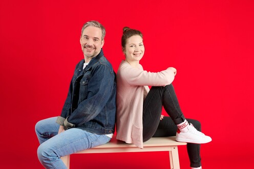 Unsere Kolleg*innen auf einer Bank Zwei unserer Kolleg*innen sitzen auf einer Holzbank vor rotem Hintergrund. Sie lächeln in die Kamera.