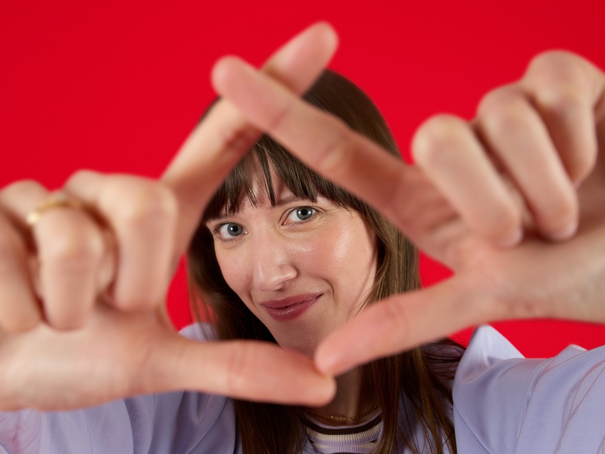 Unsere Kollegin aus Strategie ist beim Living Red Shooting. Sie trägt eine lila Jacke und steht vor rotem Hintergrund. Ihre Hände bilden ein Dreieck, durch das sie in die Kamera schaut. 