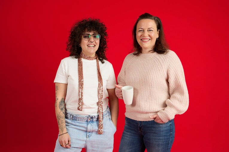 Unsere Kolleginnen lächeln in die Kamera Unsere Kolleginnen stehen vor einem roten Hintergrund und lachen in die Kamera. Sie hält eine Tasse in der Hand. Die andere trägt einen rosa Schal.