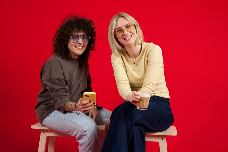 Unsere Kolleginnen sitzen beim Living Red Shooting auf einer Bank. Sie tragen beide eine Sonnenbrille und trinken einen Kaffee aus einem Glas.