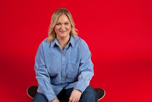 Unsere HR Managerin in Digital Learning sitzt auf einem Skateboard auf dem Boden vor rotem Hintergrund. Sie lächelt in die Kamera.