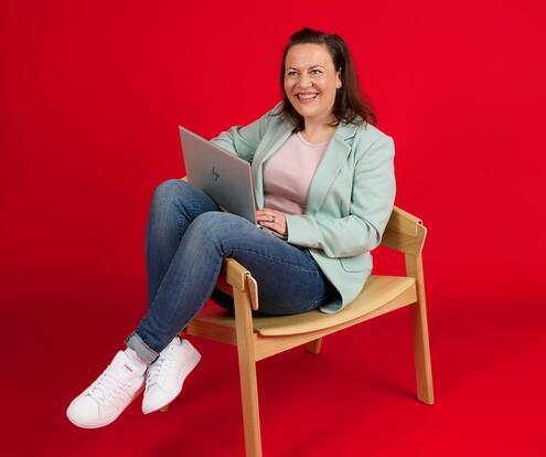 Unsere Kollegin sitzt auf einem Holzstuhl vor einem roten Hintergrund. Ihre Beine hängen entspannt über die Stuhllehne. Sie hat einen Laptop auf dem Schoß und lächelt freundlich in die Kamera. 