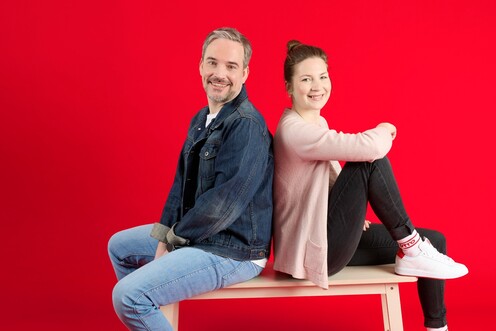 Unsere Kolleg*innen auf einer Bank Zwei unserer Kolleg*innen sitzen auf einer Holzbank vor rotem Hintergrund. Sie lächeln in die Kamera.