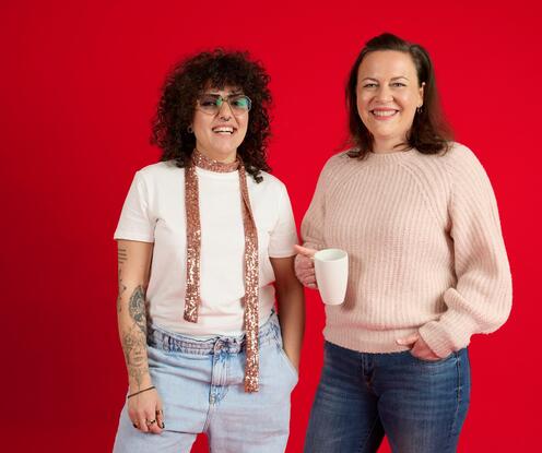 Unsere Kolleginnen stehen vor einem roten Hintergrund und lachen in die Kamera. Sie hält eine Tasse in der Hand. Die andere trägt einen rosa Schal. 
