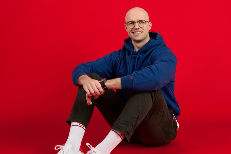 Unser Kollege sitzt lässig auf dem Boden Unser Kollege sitzt vor rotem Hintergrund und lächelt in die Kamera. Er trägt eine Brille und einen dunkelblauen Hoodie.