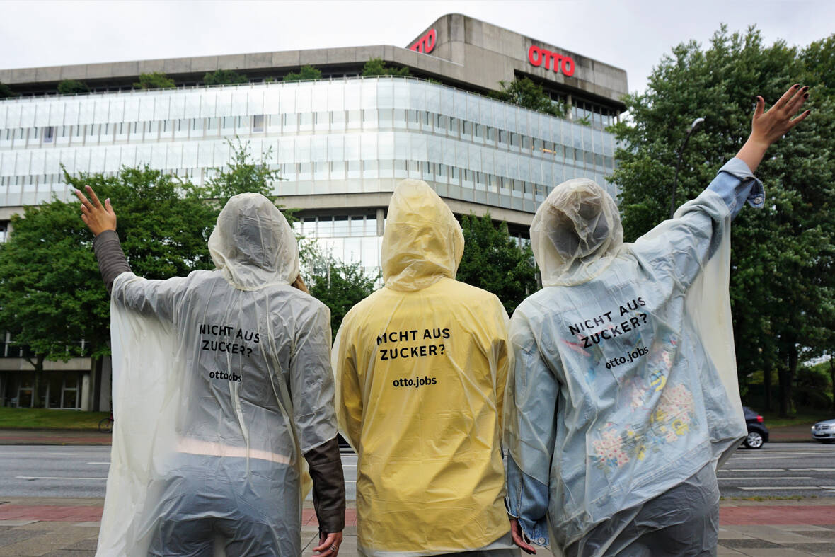 Drei Menschen mit Regenponcho stehen vor dem OTTO-Hauptgebäude. Der Himmel ist bedeckt. Auf den Regenponchos steht geschrieben "Nicht aus Zucker"
