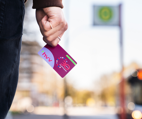 Bild einer HVV-Karte in einer Hand vor einer verschwommenen Bushaltestelle. 