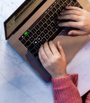 Zwei Kollegen sitzen nebeneinander am Tisch. Auf dem Tisch steht ein Glas Wasser und ein geöffneter Laptop. Einer von ihnen tippt auf dem Laptop. Zu sehen sind nur die Hände der Kollegen.