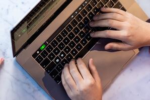 Zwei Kollegen sitzen nebeneinander am Tisch. Auf dem Tisch steht ein Glas Wasser und ein geöffneter Laptop. Einer von ihnen tippt auf dem Laptop. Zu sehen sind nur die Hände der Kollegen.