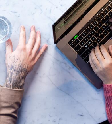 Zwei Kollegen sitzen nebeneinander am Tisch. Auf dem Tisch steht ein Glas Wasser und ein geöffneter Laptop. Einer von ihnen tippt auf dem Laptop. Zu sehen sind nur die Hände der Kollegen.