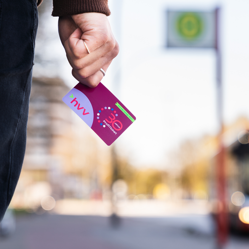 Bild einer HVV-Karte in einer Hand vor einer verschwommenen Bushaltestelle.