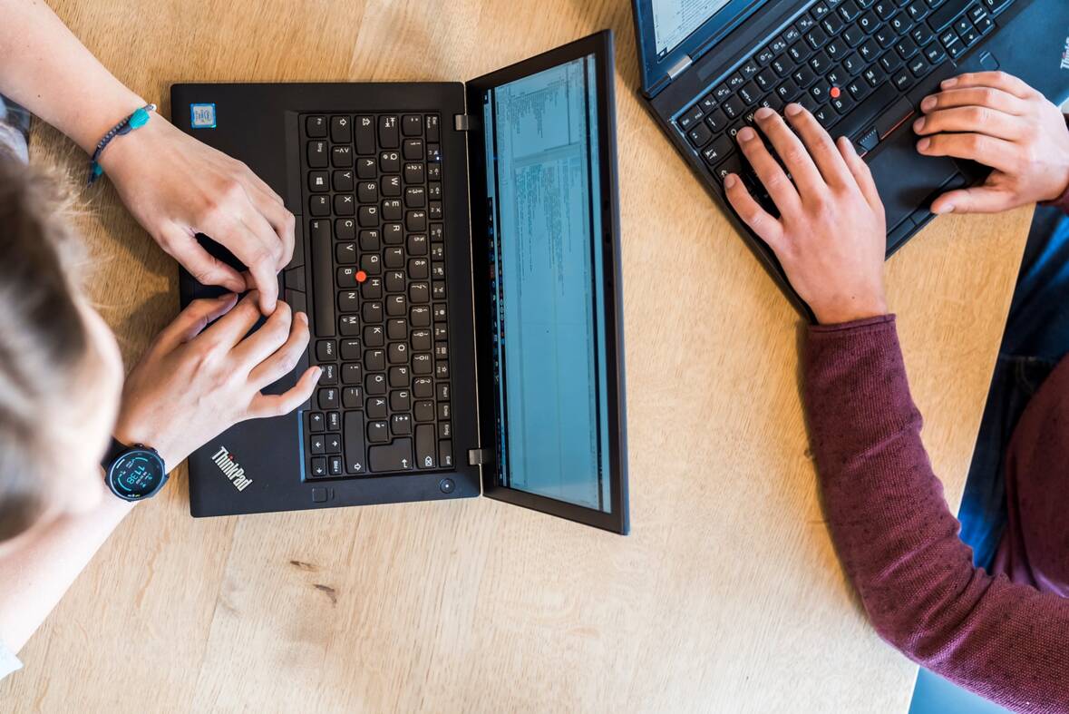 Zwei Kollegen sitzen sich mit ihren Laptopen gegenüber und tippen darauf.
