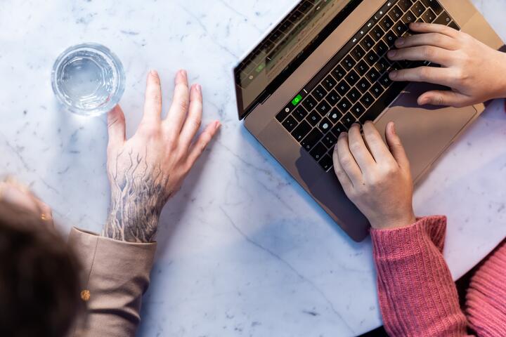 Zwei Kollegen sitzen nebeneinander am Tisch. Auf dem Tisch steht ein Glas Wasser und ein geöffneter Laptop. Einer von ihnen tippt auf dem Laptop. Zu sehen sind nur die Hände der Kollegen.