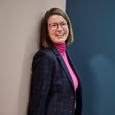 Eine Kollegin mit Brille und schulterlangen Haaren lehnt an der Wand und guckt lächelnd in die Kamera.