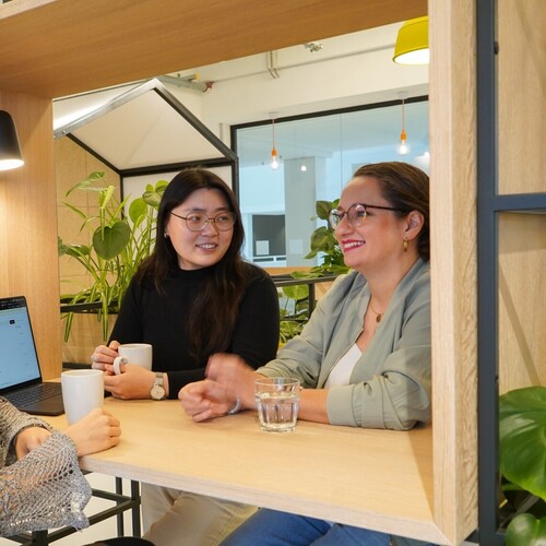 Drei Kolleginnen aus dem UX Design sitzen im Social Space an einem Tisch mit Laptop und Getränk und unterhalten sich. Im Hintergrund sind mehrere Pflanzen.