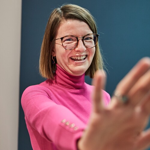 Eine Kollegin mit Brille und schulterlangen Haaren lächelt in die Kamera.