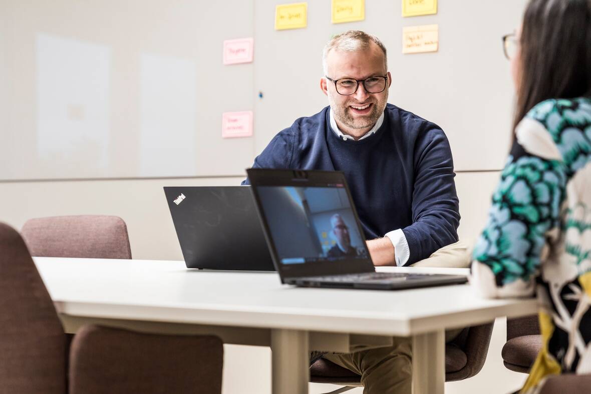 Zwei Kollegen sitzen zusammen am Tisch in einem Meetingraum. Auf den Laptops vor ihnen lässt sich erkennen, dass noch weitere Kollegen gerade am Meeting teilnehmen. An dem Whiteboard an der Wand hängen bereits Haftnotizzettel.