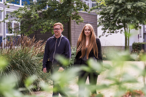 Zwei Mitarbeitende gehen auf dem gepflasterten Weg des Campus Boulevards entlang. An der Seite des Weges sind viele Grünflächen mit Büschen und Bäumen angelegt und im Hintergrund steht ein Gebäude