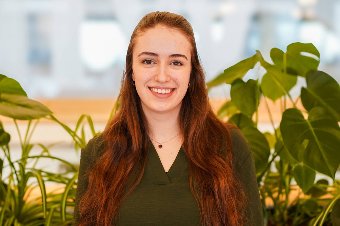 Unsere Duale Studentin lächelt in die Kamera und im Hintergrund sind mehrere Pflanzen zu erkennen. Sie befindet sich in einem unserer Social Spaces. 