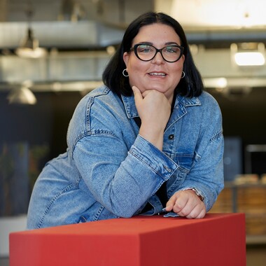 Unsere Kollegin arbeitet im Callcenter von OTTO, trägt eine Brille und lehnt lächelnd auf einem roten Hochtisch.