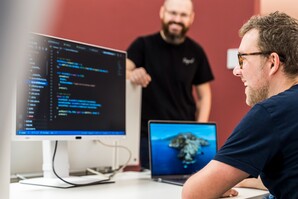 Ein IT-Mitarbeiter sitzt am Schreibtisch und guckt auf den Code, der auf dem Computerbildschirm angezeigt wird. Ein anderer Kollege steht freundlich daneben.