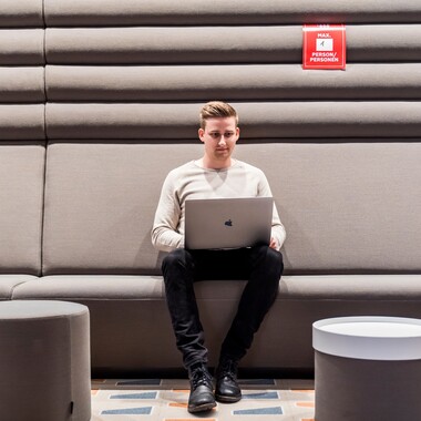 Ein junger Kollege, der als Digital Analyst arbeitet, sitzt mit seinem Laptop auf einem grauen Sofa in einem unserer Meetingräume und ist ganz konzentriert.