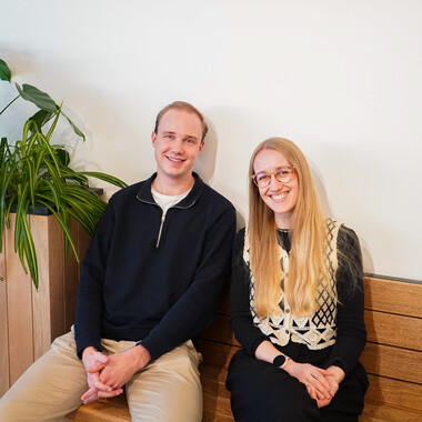 Zwei Azubis bei OTTO befinden sich im Social Space und sitzen auf einer Holzbank mit Pflanzen im Hintergrund.