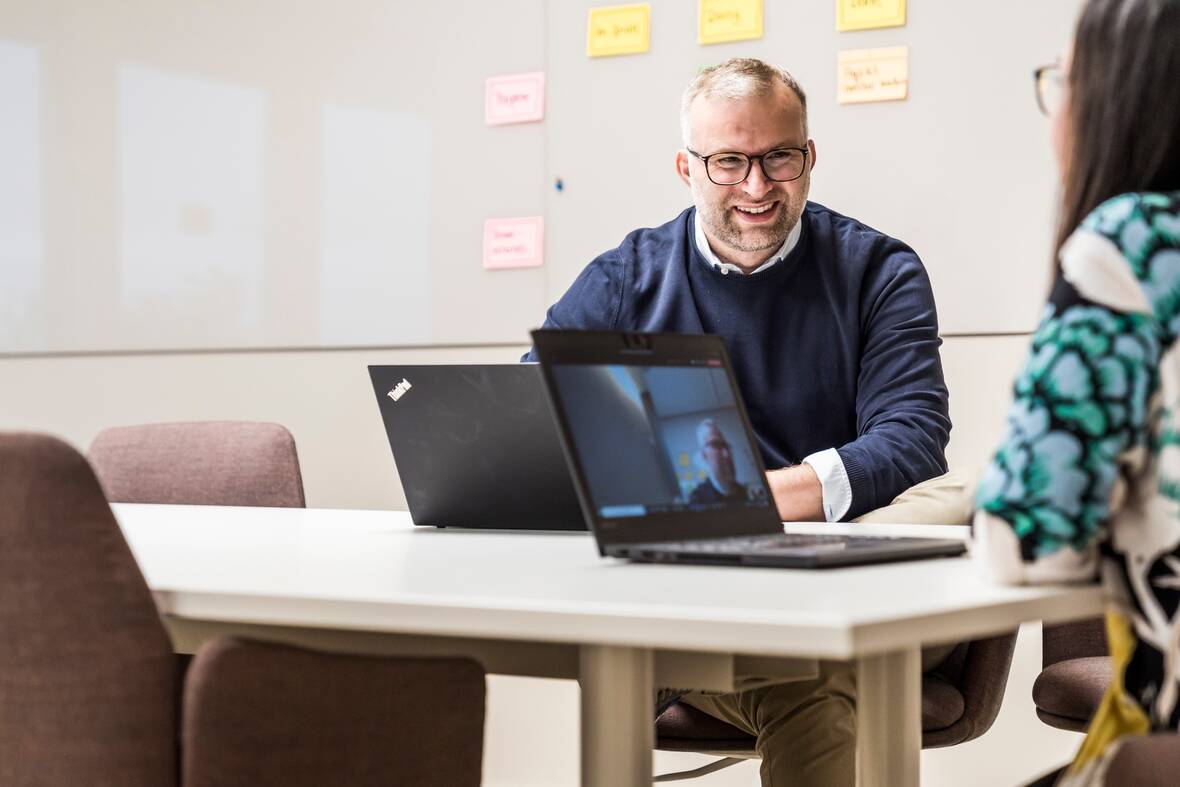 Zwei Kollegen sitzen zusammen am Tisch in einem Meetingraum. Auf den Laptops vor ihnen lässt sich erkennen, dass noch weitere Kollegen gerade am Meeting teilnehmen. An dem Whiteboard an der Wand hängen bereits Haftnotizzettel.