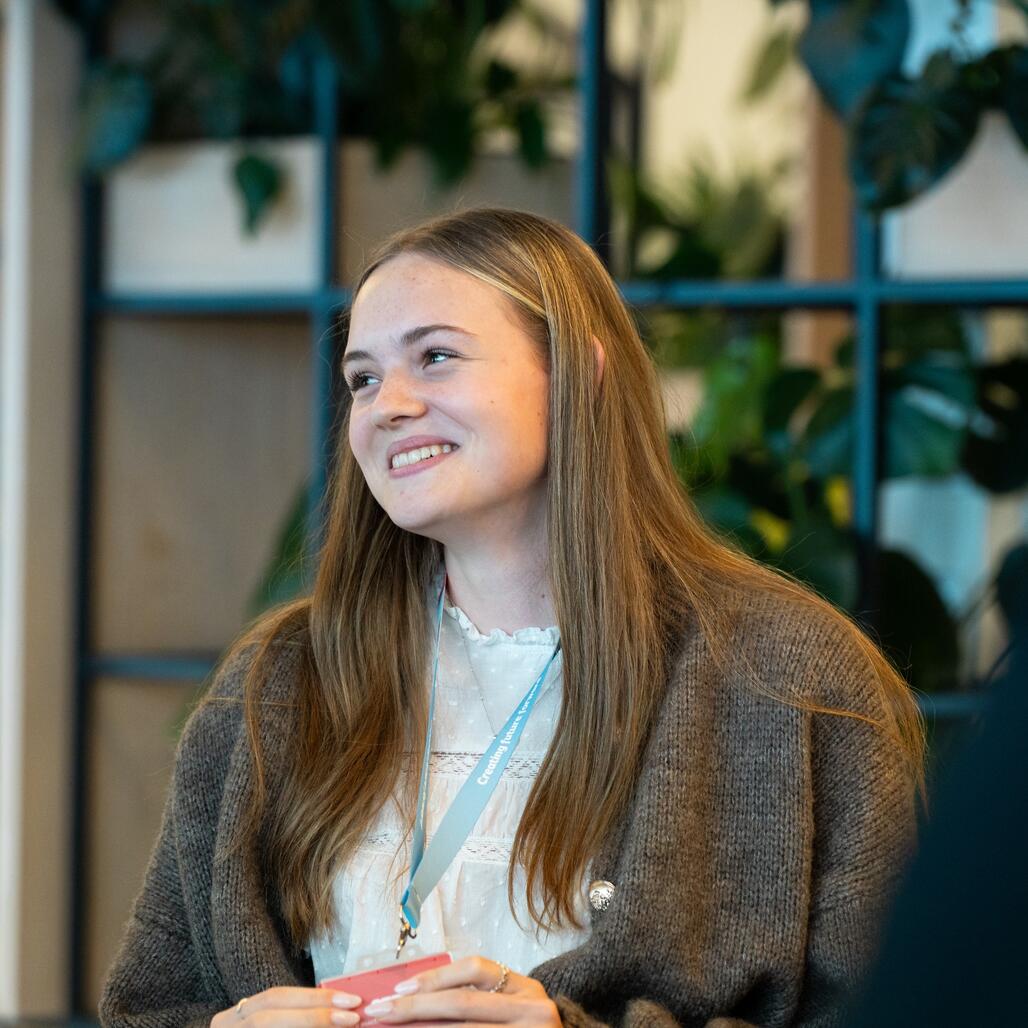 Azubi aus dem Groß- & Außenhandel lächelt zur Seite Bei den Starter Days 2025 sitzt die Auszubildende im Bereich Groß- und Außenhandel auf einem Stuhl, lächelt zur Seite und hört aufmerksam zu.