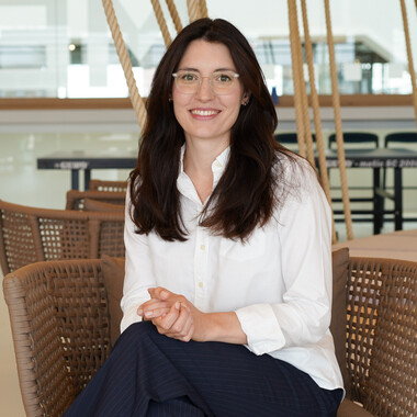 Unsere Kollegin aus dem Sales Marketing sitzt im Social Space auf dem Stuhl. Sie trägt ein weißes Hemd und eine dunkle Hose. Sie lächelt in die Kamera.