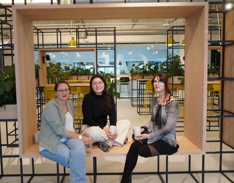 Drei Kolleginnen aus dem Bereich UX-Design sind im Social Space in einer lockeren Haltung sitzen auf einem Holzgerüst