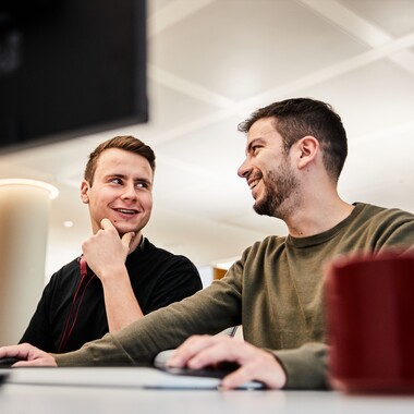 Zwei Kollegen aus Tech sitzen gemeinsam vor dem PC und unterhalten sich lächelnd.