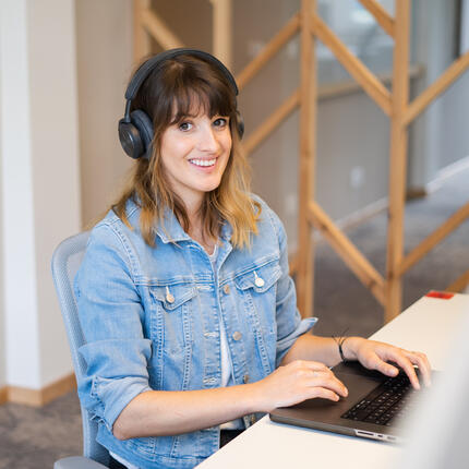 Junior Software-Entwicklerin sitzt am Computer Junior Software-Entwicklerin sitzt am Schreibtisch mit Headset und tippt am Computer und lächelt in die Kamera.
